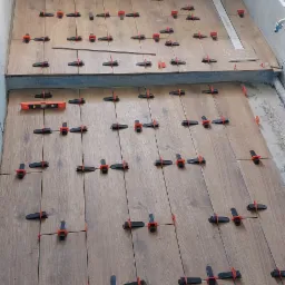 Układanie płytek imitujących drewno w łazience, z użyciem poziomicy i klipsów dystansowych, widok z góry na podłogę i fragment ściany w surowym stanie.