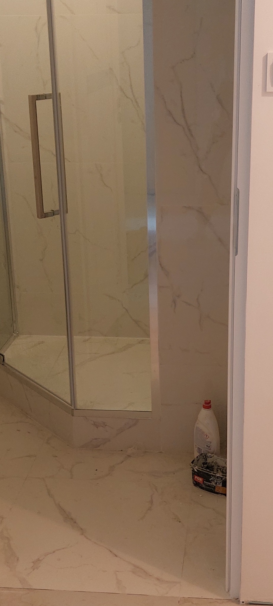 Wykończona kabina prysznicowa z marmurowymi płytkami na ścianach i podłodze. Widoczny podniesiony brodzik z odpływem liniowym, butelka i wiadro.