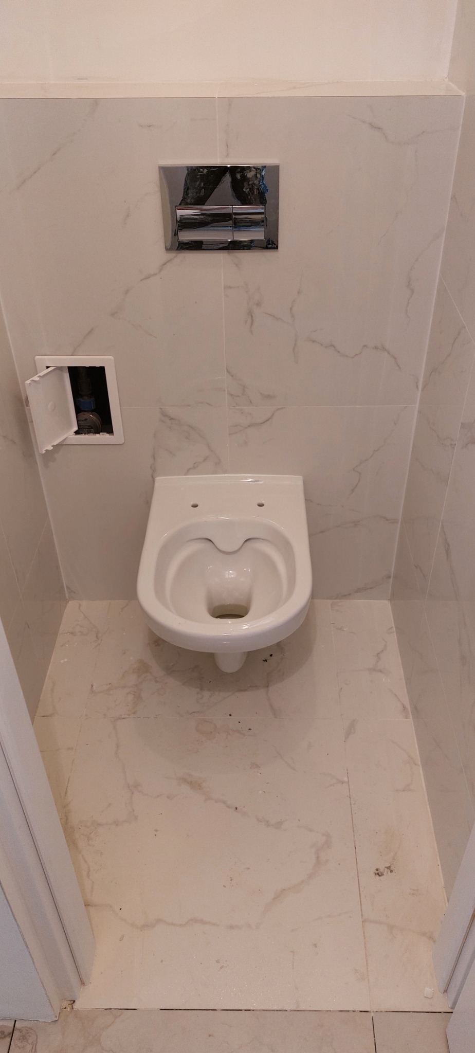 Nowoczesna toaleta podwieszana w łazience wykończonej marmurowymi płytkami, z chromowanym przyciskiem spłukującym i otwartą klapką rewizyjną.