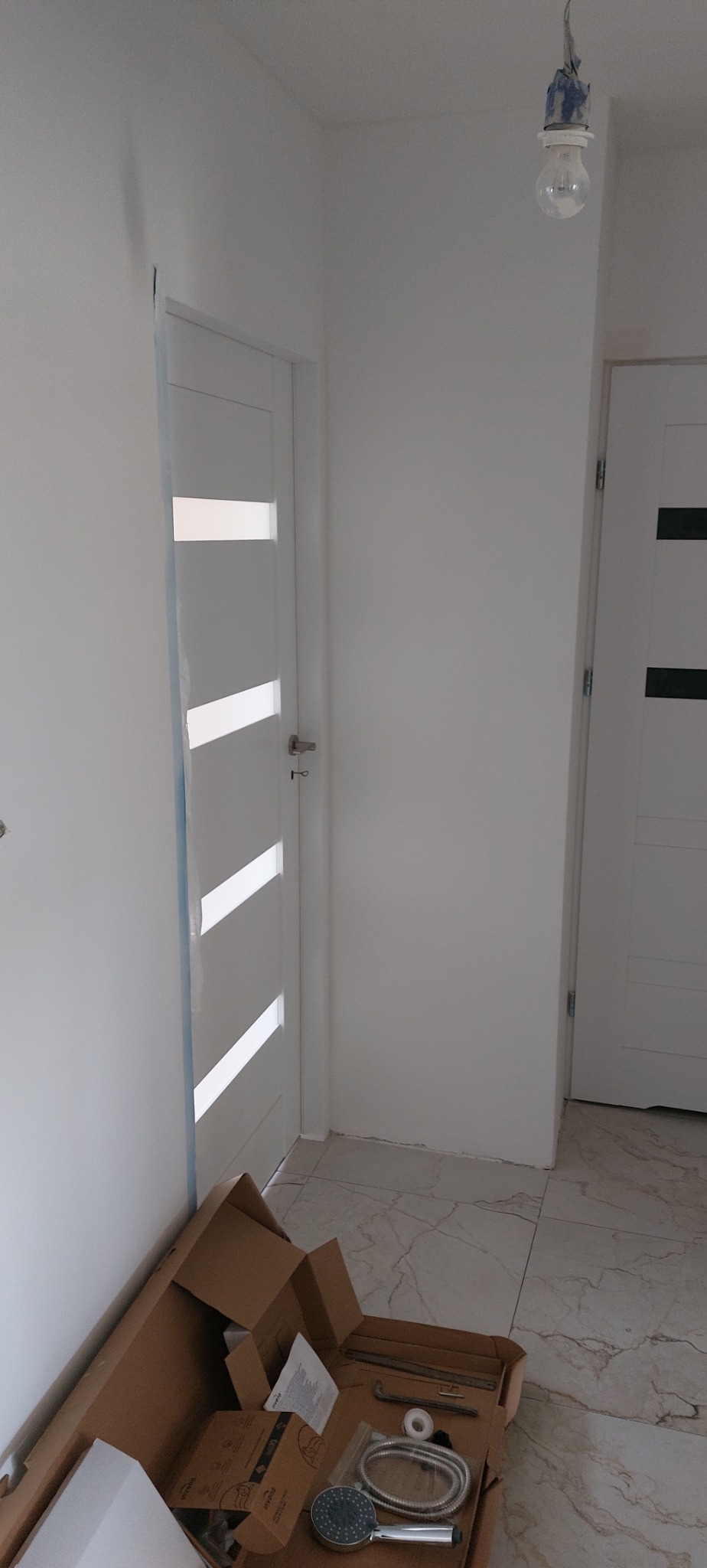 Białe drzwi wewnętrzne z przeszkleniami w trakcie montażu, widoczna kartonowa skrzynka z elementami armatury łazienkowej na marmurowej posadzce.