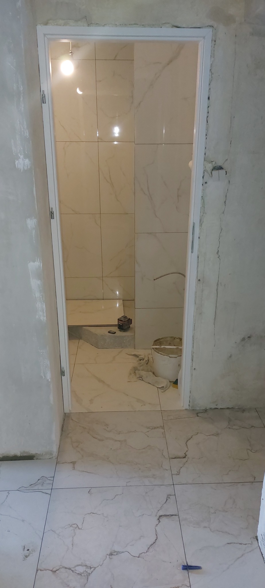 Wykończona łazienka z marmurowymi płytkami, widoczny brodzik i białe drzwi. Na podłodze leży narzędzie glazurnicze, a w rogu stoi wiadro z zaprawą.