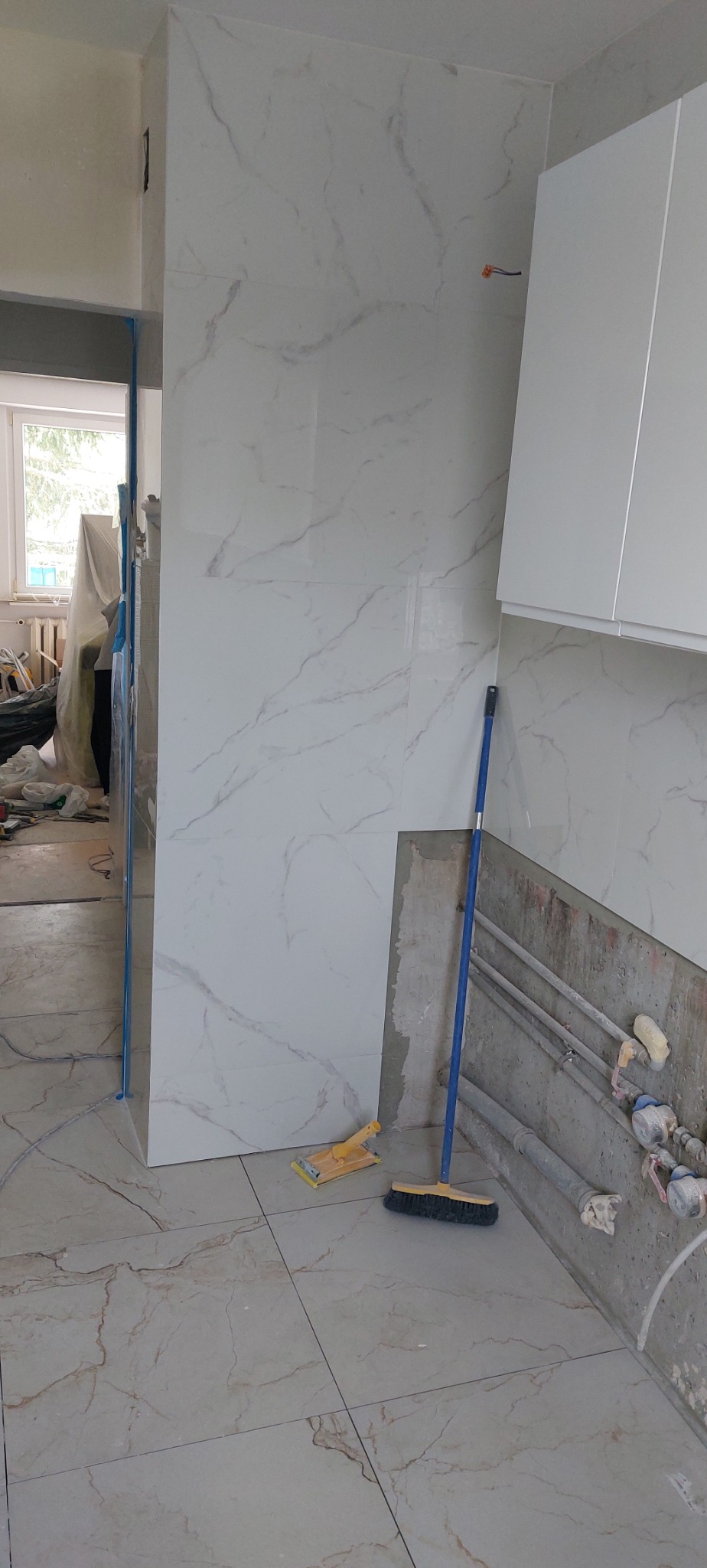 Układanie płytek imitujących marmur w łazience. Widoczne narzędzia glazurnicze i fragment instalacji hydraulicznej. Prace wykończeniowe w toku.