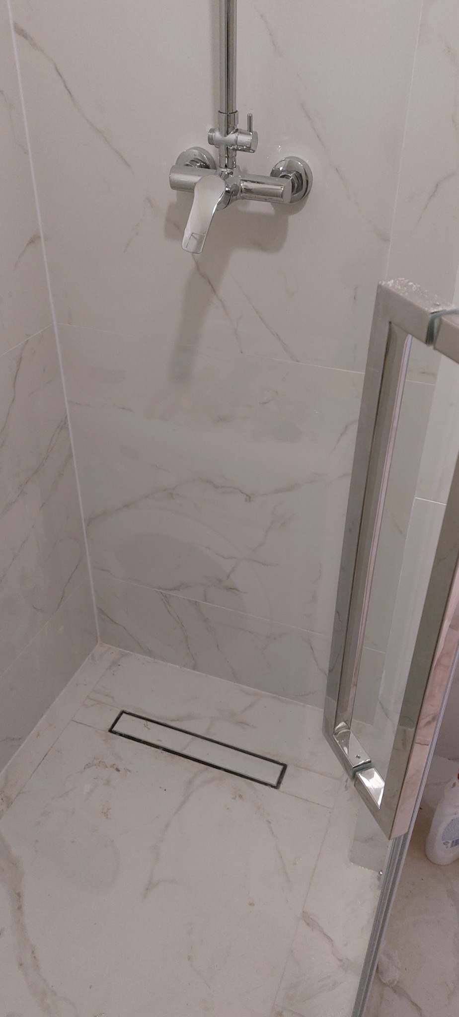 Wykończona kabina prysznicowa z odpływem liniowym i armaturą w kolorze chromu. Białe płytki imitujące marmur na ścianach i podłodze. Częściowo widoczne szklane drzwi.