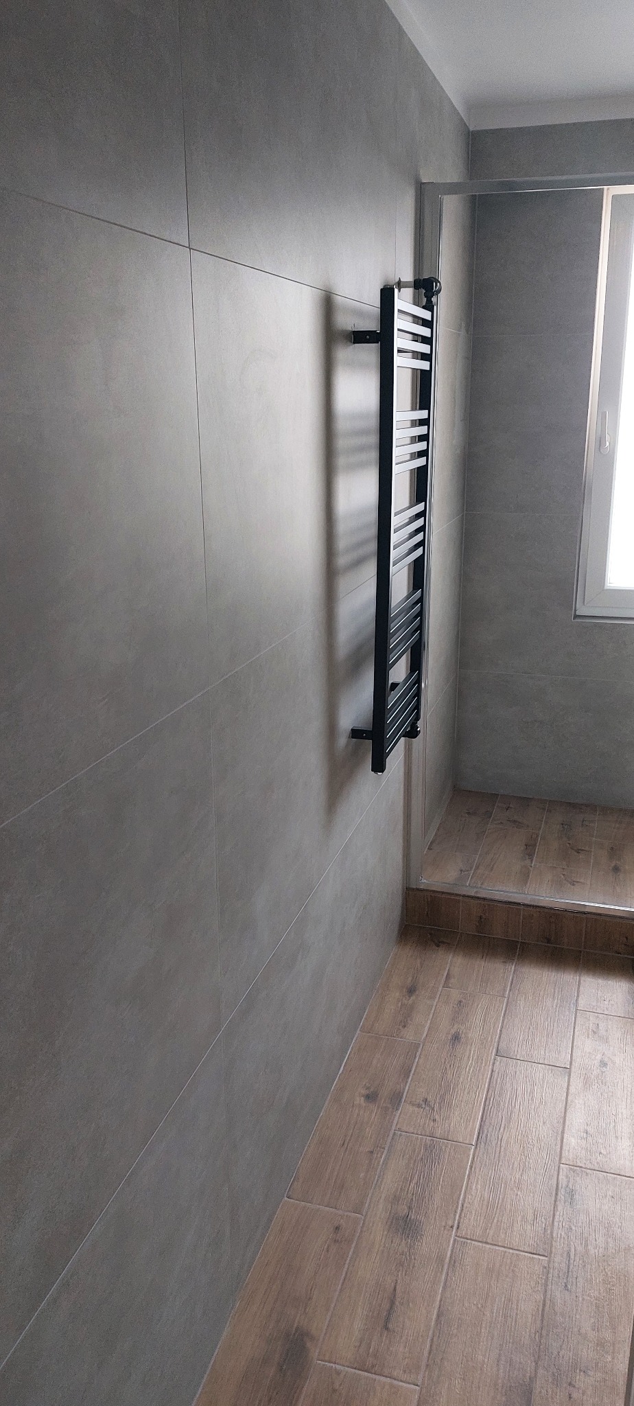 Minimalistyczna łazienka po remoncie: szare, duże płytki na ścianach, drewnopodobne płytki na podłodze, czarny grzejnik drabinkowy, kabina prysznicowa.