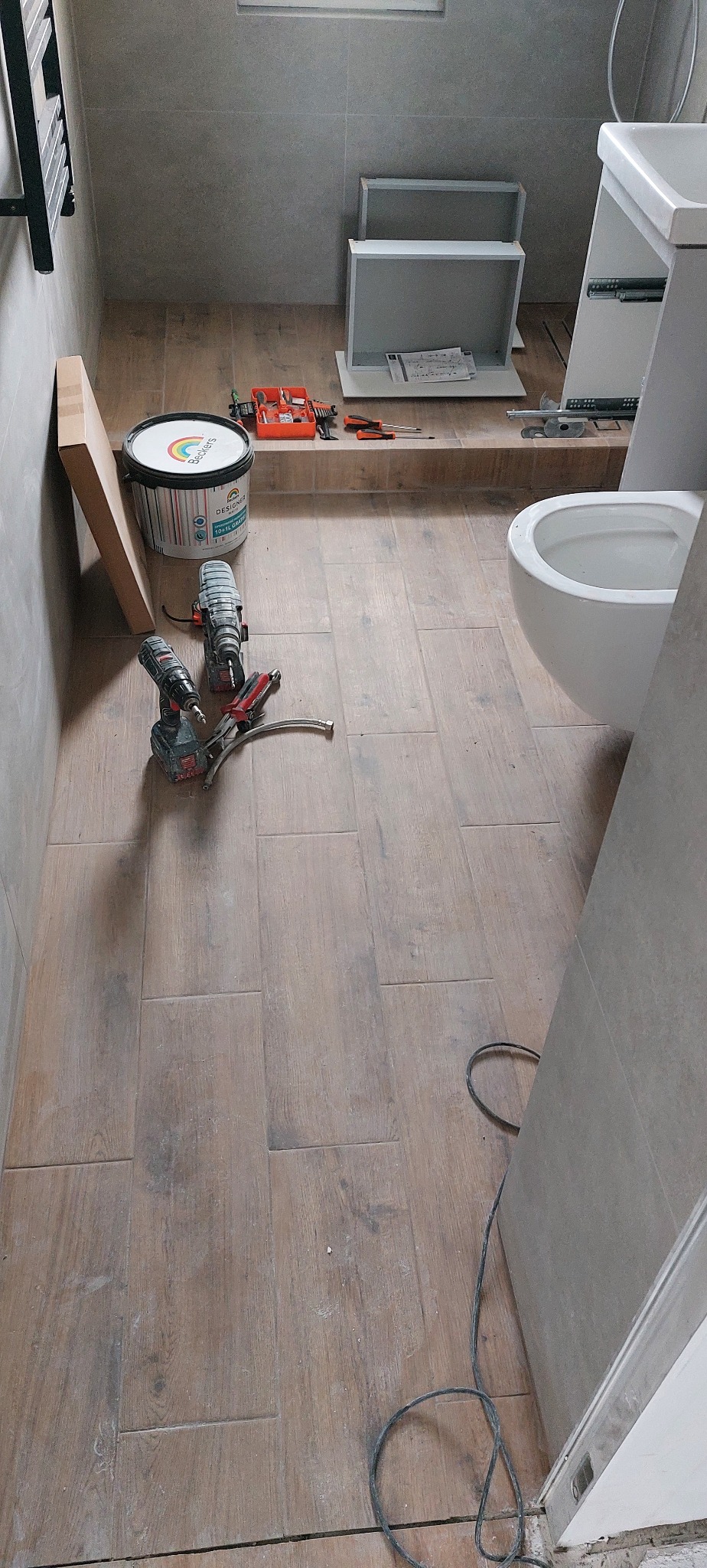 Łazienka w trakcie prac: ułożone płytki imitujące drewno, narzędzia, wiadro z farbą, elementy szafki pod umywalkę i toaleta w tle. Widok z góry.