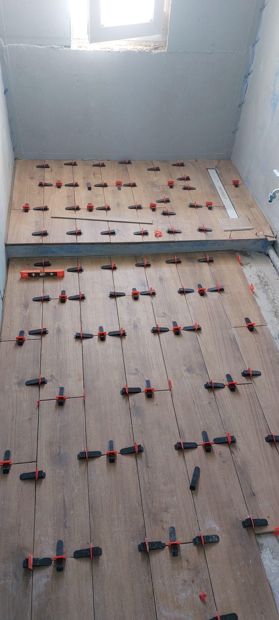 Układanie płytek imitujących drewno w łazience, z użyciem poziomicy i klipsów dystansowych, widok z góry na podłogę i fragment ściany w surowym stanie.