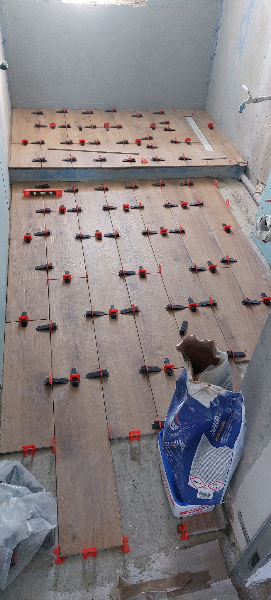 Układanie podłogi z płytek imitujących drewno z użyciem klipsów poziomujących. Widoczny worek z klejem. Etap prac w trakcie.