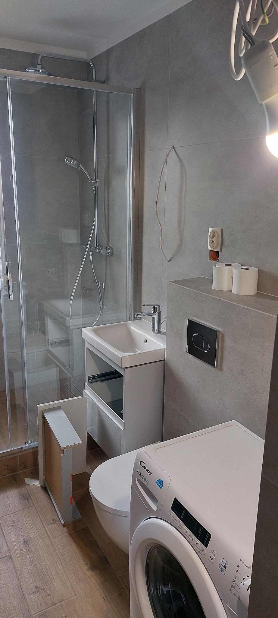 Szara łazienka w trakcie wykańczania: kabina prysznicowa, umywalka z otwartą szafką, toaleta i pralka Candy. Widoczne niedokończone detale.