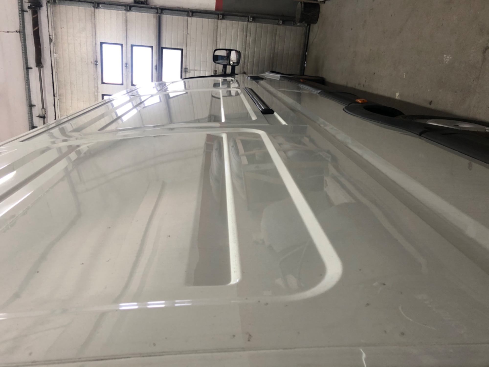 Biały dach samochodu dostawczego widziany pod kątem, z widocznymi elementami konstrukcyjnymi dachu, lusterkiem wstecznym i fragmentem dachu nad kabiną kierowcy, na tle bramy garażowej.