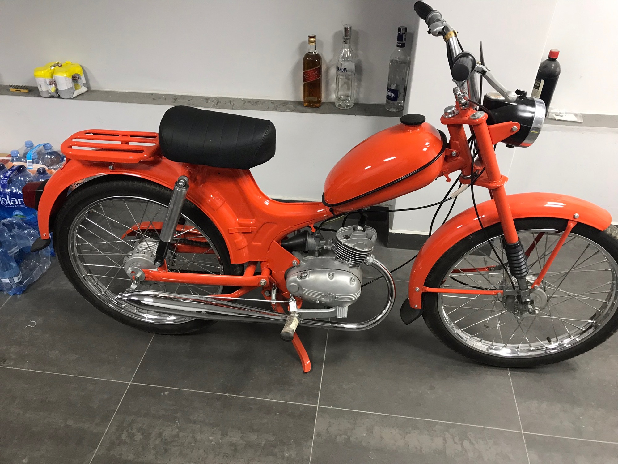 Odrestaurowany motorower Romet TYP 100 w jaskrawym pomarańczowym lakierze, widoczny na tle ściany z butelkami alkoholu i zapasem wody mineralnej.