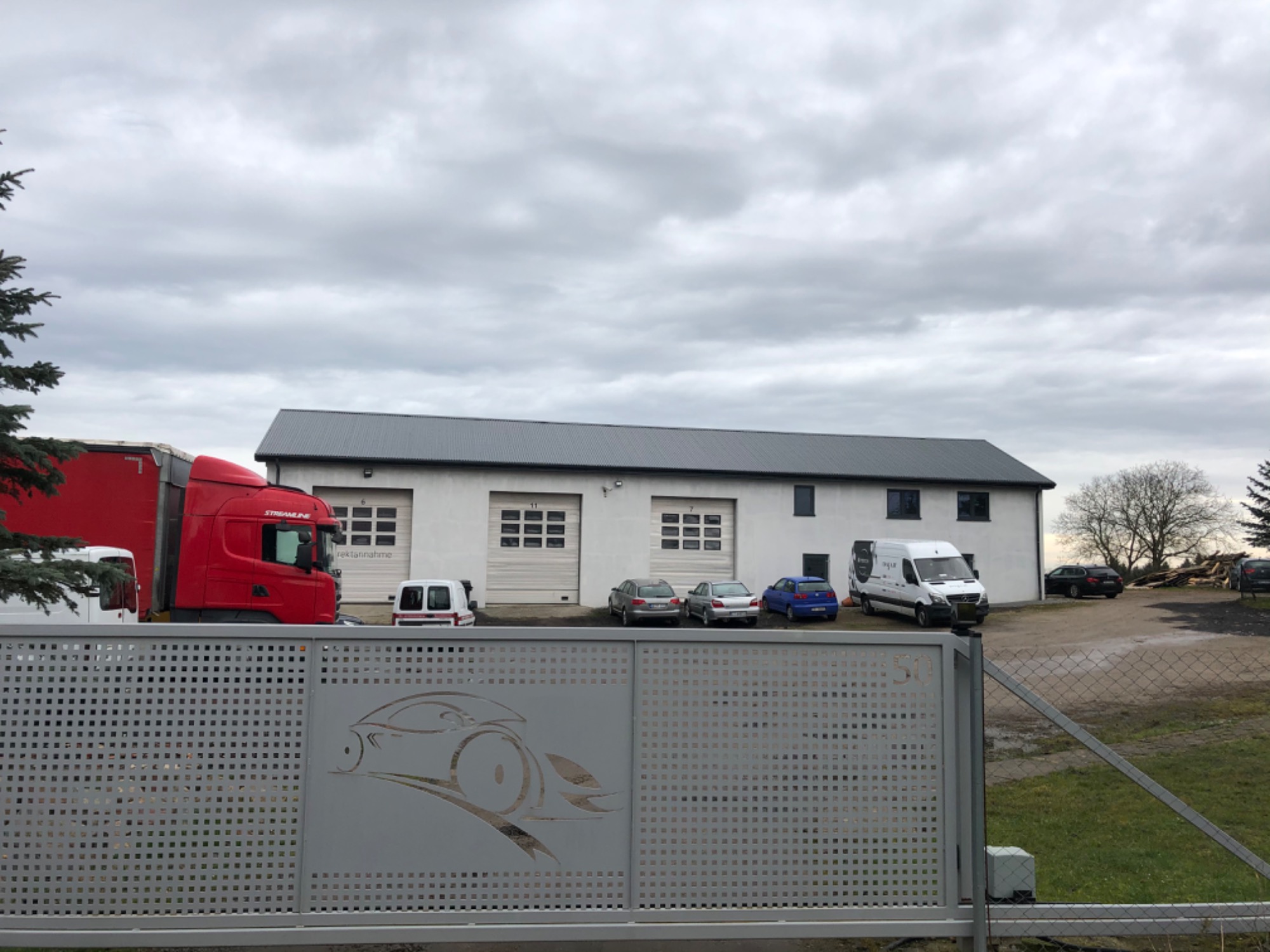 Biały budynek warsztatu samochodowego z trzema bramami garażowymi, widoczny zza metalowej bramy z wyciętym wzorem samochodu, z zaparkowanymi samochodami osobowymi, dostawczymi i ciężarowymi na placu.