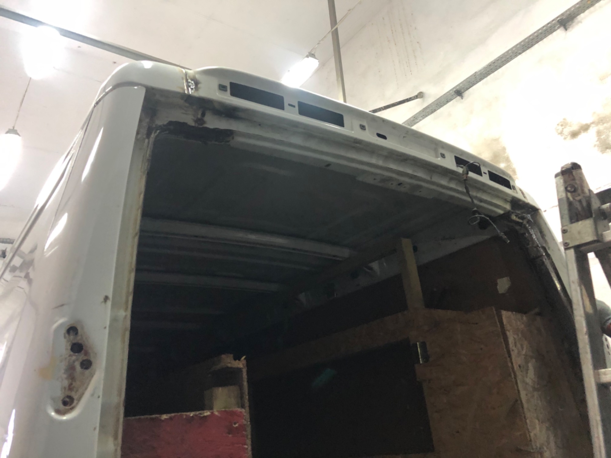 Uszkodzona tylna część białego samochodu dostawczego w warsztacie, widoczne ślady korozji i napraw blacharskich, drewniana zabudowa wnętrza, oświetlenie warsztatowe.