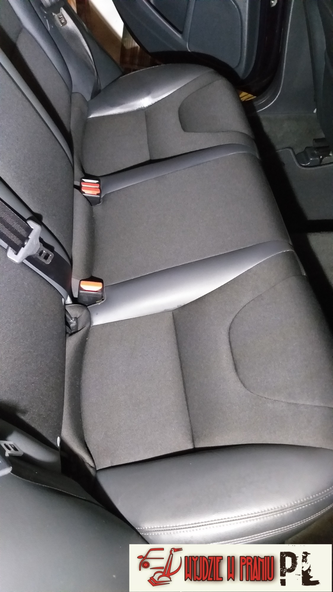 Czarne, materiałowe tylne siedzenie samochodowe z pasami bezpieczeństwa, widoczne detale przeszyć i logo firmy czyszczącej w prawym dolnym rogu.