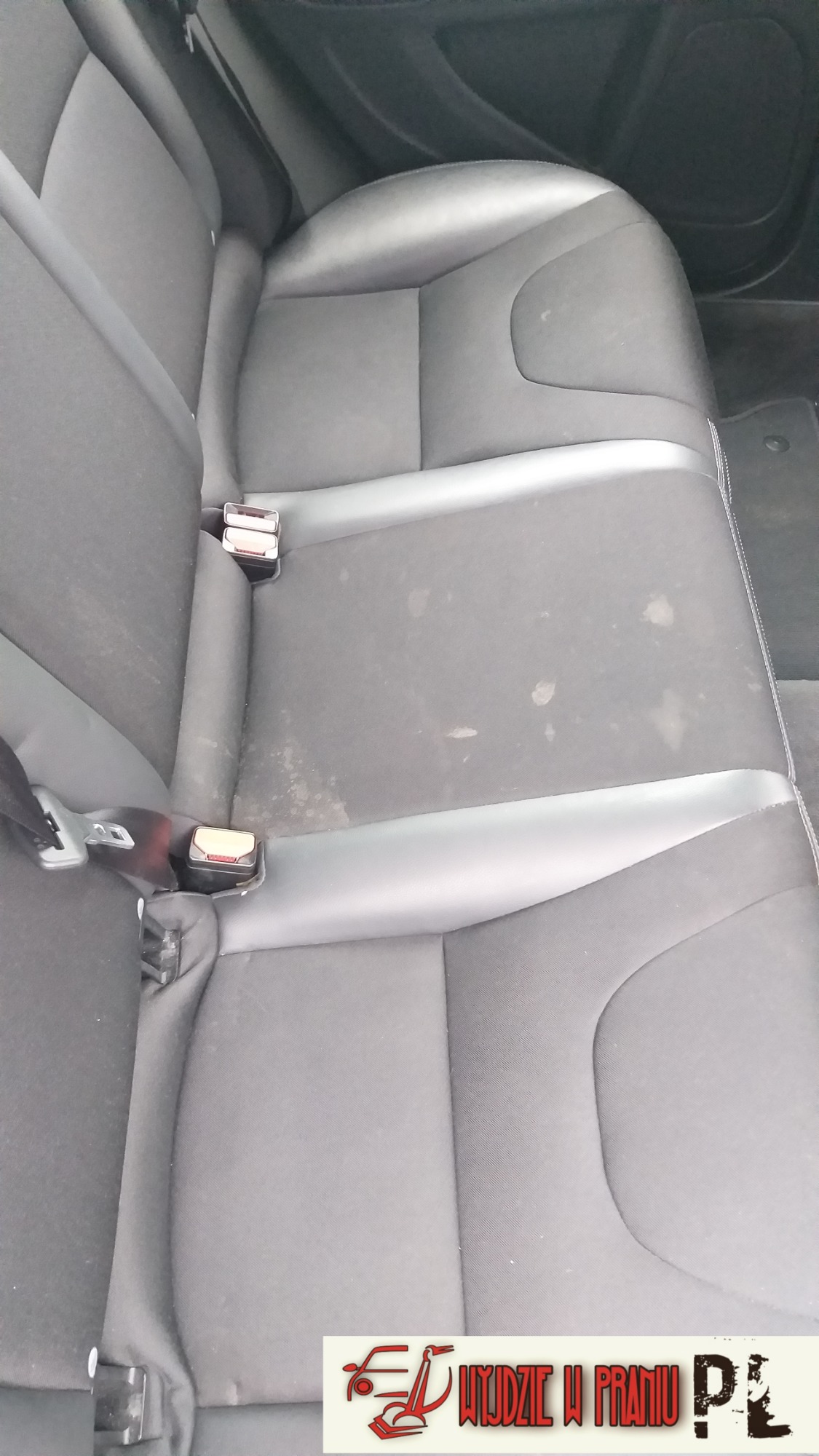 Tylna kanapa samochodu z widocznymi plamami na tapicerce, przed czyszczeniem, zapięcia pasów bezpieczeństwa widoczne.