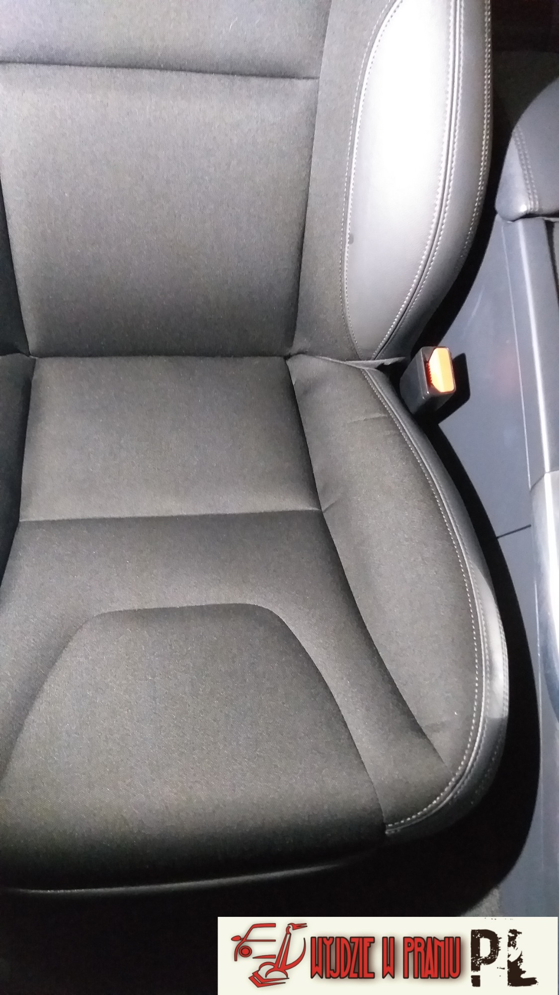Szare, materiałowe siedzenie samochodowe z widocznym zapięciem pasa bezpieczeństwa i fragmentem plastikowego panelu bocznego, na dole widoczne logo firmy.