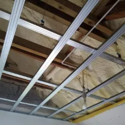 Metalowy stelaż sufitowy zamontowany pod drewnianą konstrukcją z izolacją piankową, widoczna surowa żarówka na kablu.