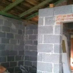 Wnętrze w trakcie budowy: ściany z szarego bloczka betonowego, fragment ściany z czerwonej cegły nad otworem drzwiowym, widoczne elementy konstrukcji dachu i sylwetki dwóch osób w tle.