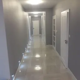 Długi, jasny korytarz z szarymi ścianami, białymi drzwiami i podświetlaną podłogą z płytek imitujących marmur. Małe punkty świetlne wzdłuż ściany dodają nowoczesnego charakteru.