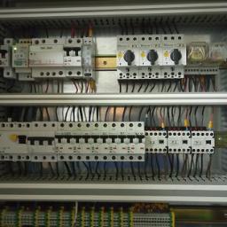Timed - Rozdzielnica elektryczna z licznymi zabezpieczeniami i okablowaniem, zainstalowana w Warszawie. Widoczne bezpieczniki, styczniki i listwy zaciskowe wewnątrz metalowej obudowy.