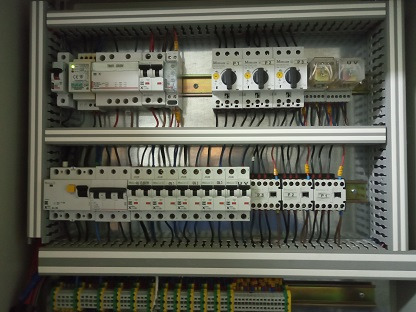 Rozdzielnica elektryczna z licznymi zabezpieczeniami i okablowaniem, zainstalowana w Warszawie. Widoczne bezpieczniki, styczniki i listwy zaciskowe wewnątrz metalowej obudowy.