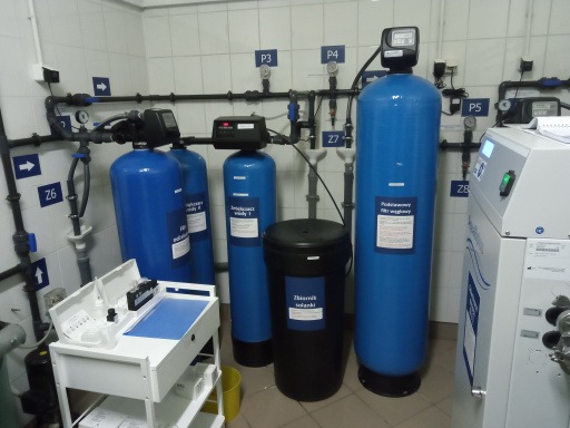 Kompleksowa instalacja filtrów do uzdatniania wody w pomieszczeniu technicznym z białymi kafelkami, widoczne niebieskie zbiorniki filtracyjne, czarny zbiornik solanki, orurowanie z oznaczeniami...