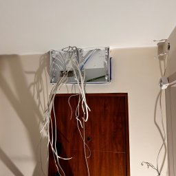 USŁUGI BUDOWLANE TOMASZ SZCZAWIK - Rozdzielnica elektryczna z wystającymi przewodami na ścianie nad drewnianymi drzwiami. Widoczny fragment drabiny. Etap prac instalacyjnych w jasnym wnętrzu.