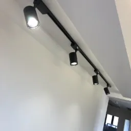 Cztery czarne reflektory punktowe LED zamontowane na szynie sufitowej w nowoczesnym wnętrzu, z widocznym fragmentem korytarza i schodów w tle.