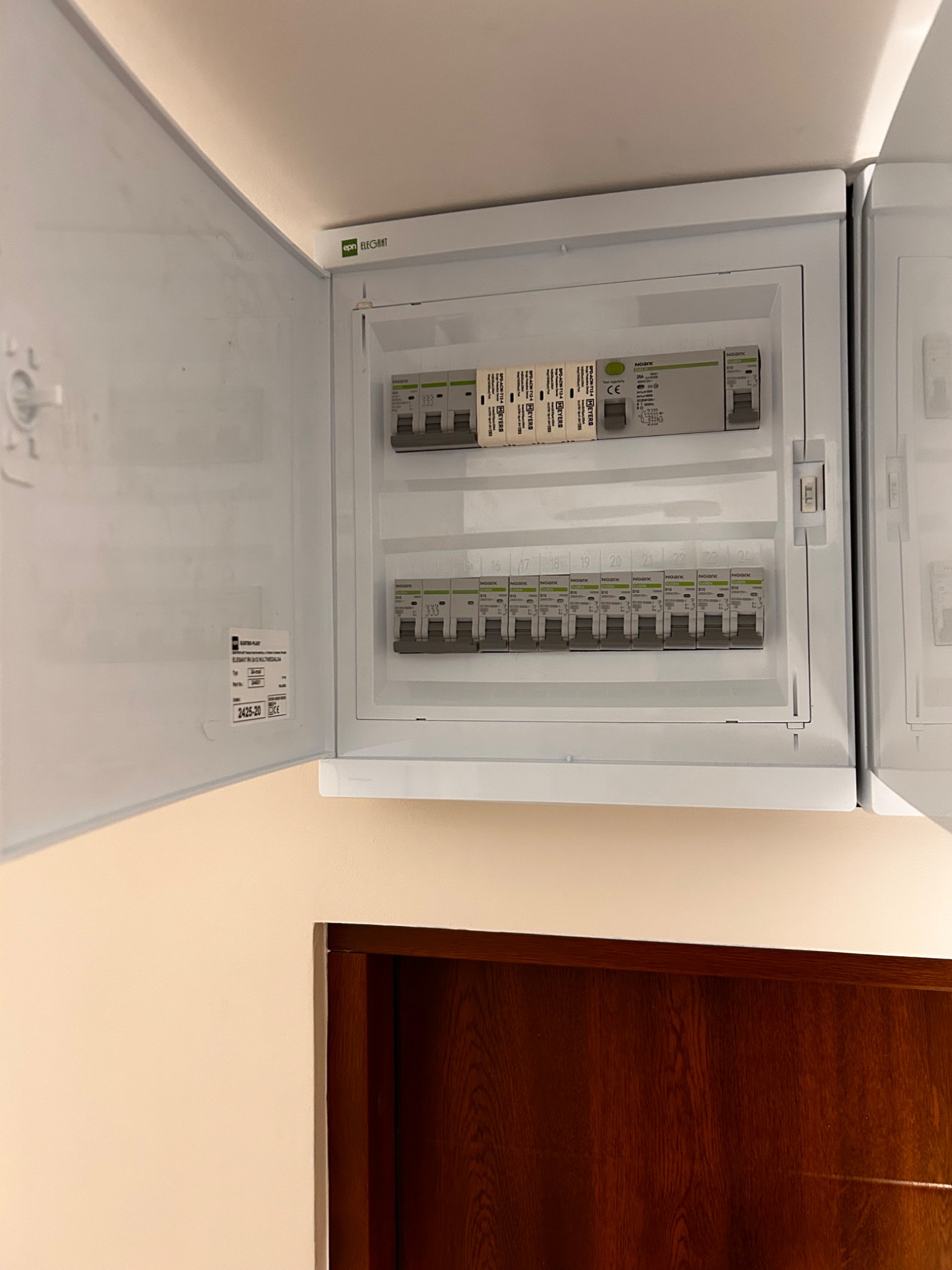 Otwarta, biała skrzynka rozdzielcza z bezpiecznikami NOARK, zamontowana na ścianie nad ciemnobrązowymi drzwiami. Widoczne oznaczenia i numery na bezpiecznikach.