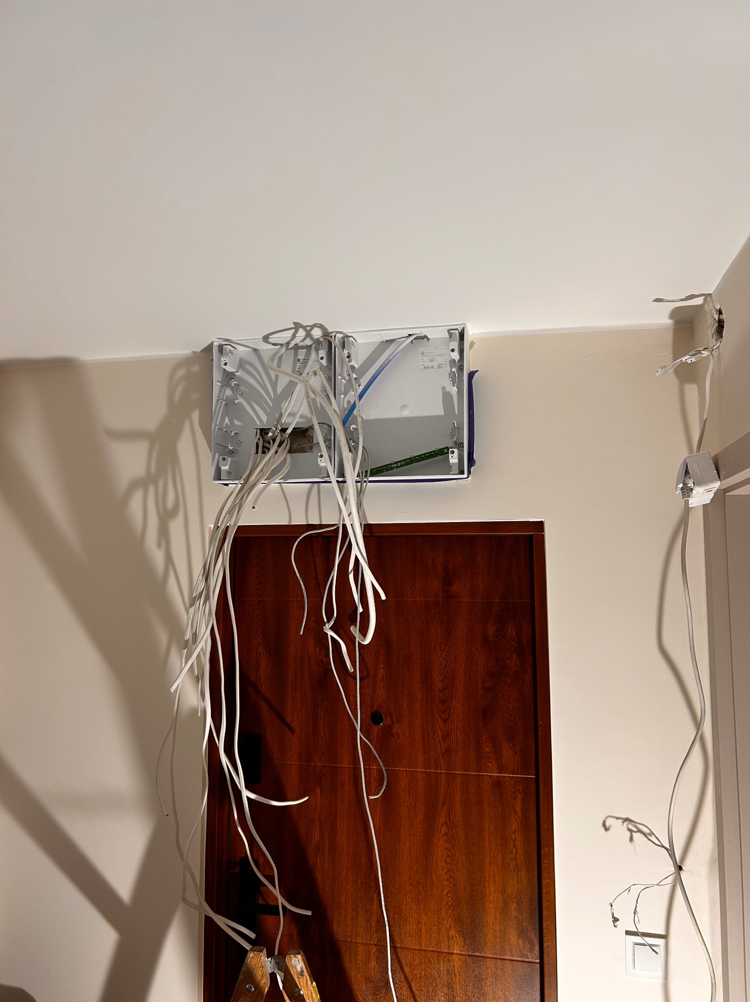 Rozdzielnica elektryczna z wystającymi przewodami na ścianie nad drewnianymi drzwiami. Widoczny fragment drabiny. Etap prac instalacyjnych w jasnym wnętrzu.
