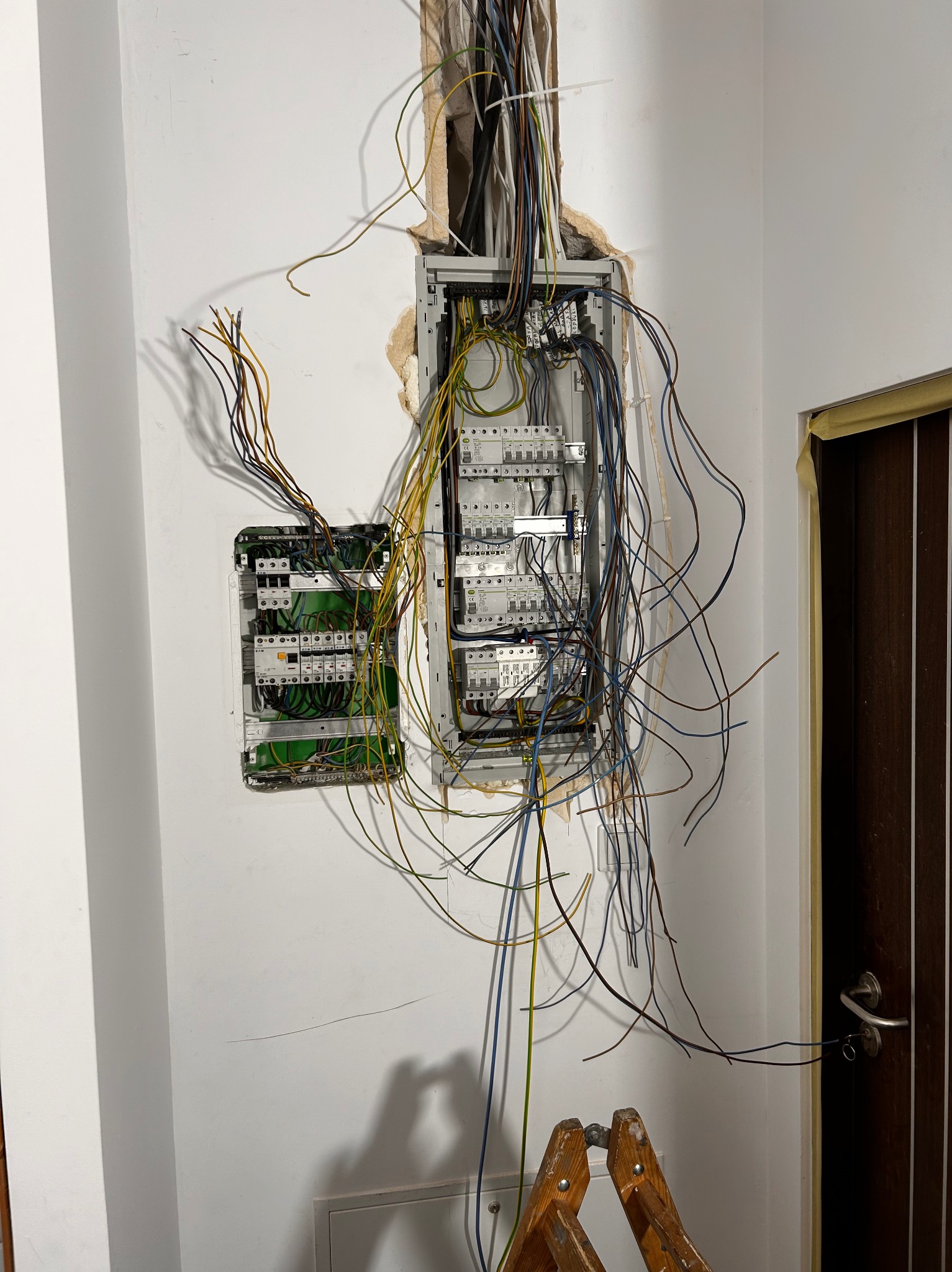 Dwa panele elektryczne w trakcie montażu na białej ścianie, widoczne liczne przewody, drabina w dolnej części kadru, obok ościeżnica drzwiowa z taśmą malarską.