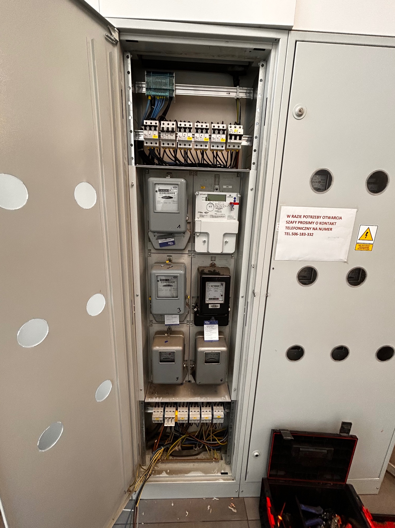 Otwarta szafa elektryczna z zamontowanymi licznikami i bezpiecznikami. Widoczna skrzynka z narzędziami elektrycznymi. Tabliczka informacyjna z numerem telefonu.