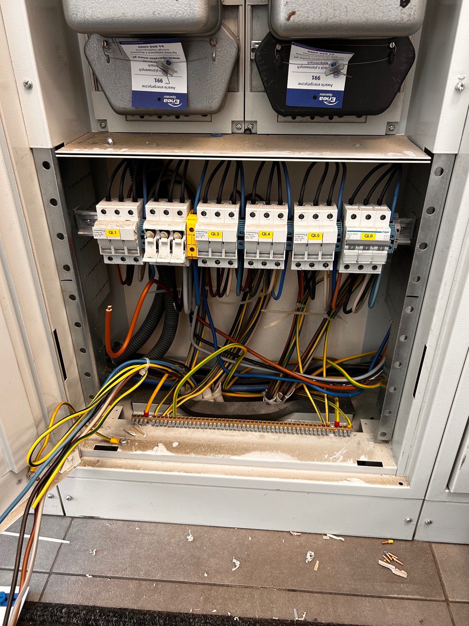 Otwarta skrzynka elektryczna z bezpiecznikami i kolorowymi przewodami. Widoczne oznaczenia QL1-QL6 na bezpiecznikach. U dołu listwa zaciskowa i fragment podłogi z resztkami po pracy.