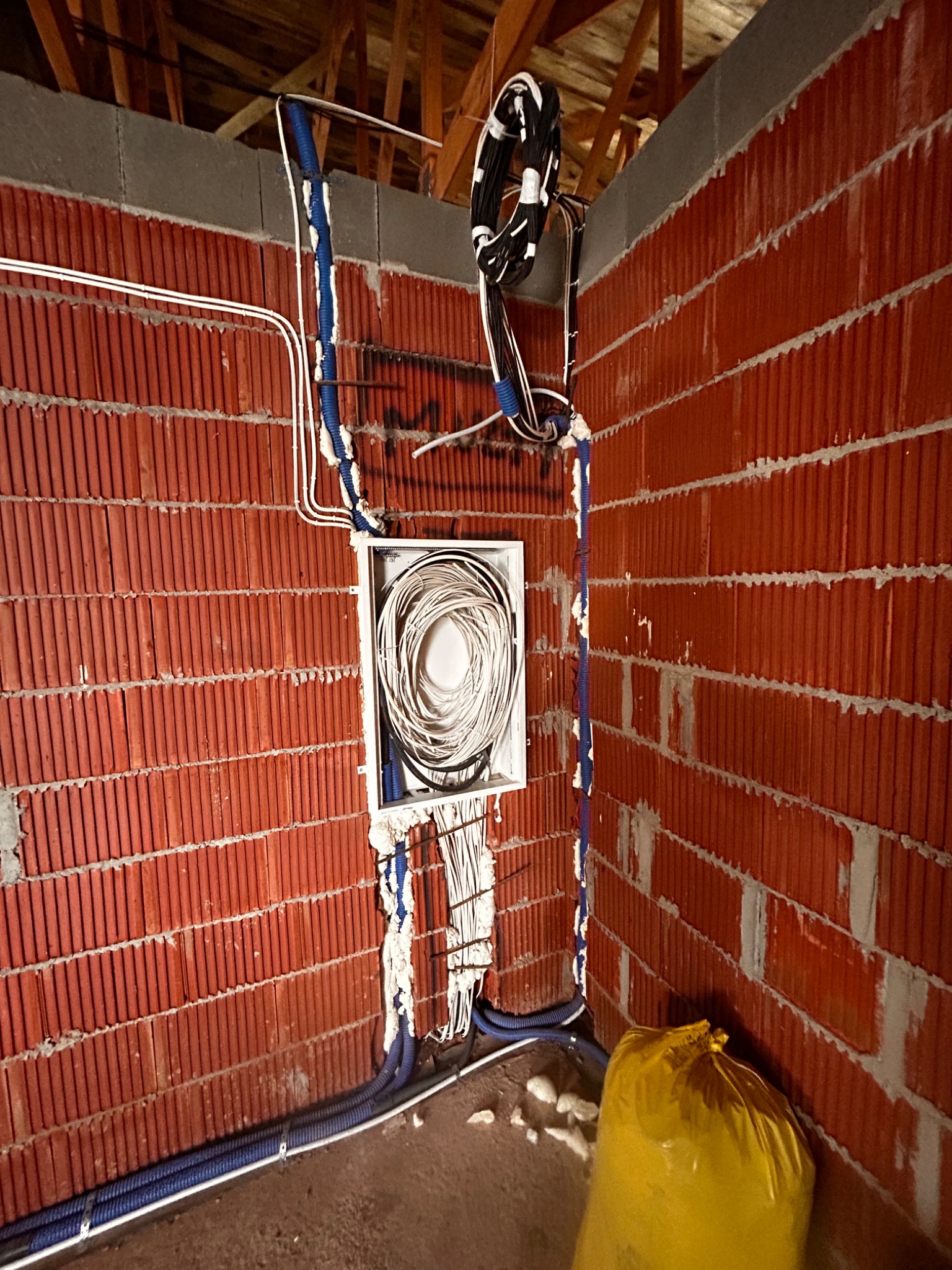 Rozprowadzenie przewodów w peszlach na ceglanej ścianie, widoczna puszka instalacyjna z kablami. Elementy instalacji zabezpieczone pianką montażową. W tle drewniana konstrukcja dachu.