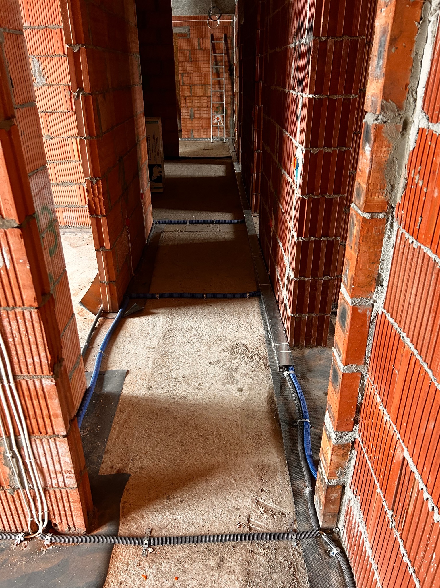 Wnętrze w trakcie budowy z widocznymi przewodami w peszlach, ułożonymi wzdłuż ścian z czerwonej cegły. Widoczne kable elektryczne w białych rurkach. Surowy stan.