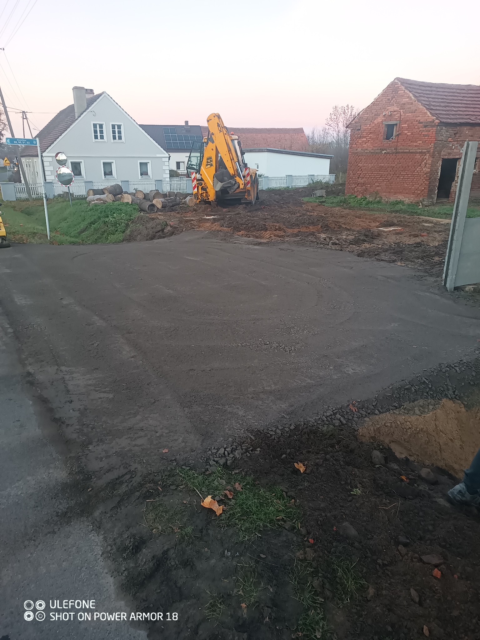 Żółta koparko-ładowarka na utwardzonym terenie, w tle domy i drzewa. Widoczne ślady opon. Utwardzona droga z gruzem na obrzeżach. Krajobraz wiejski.