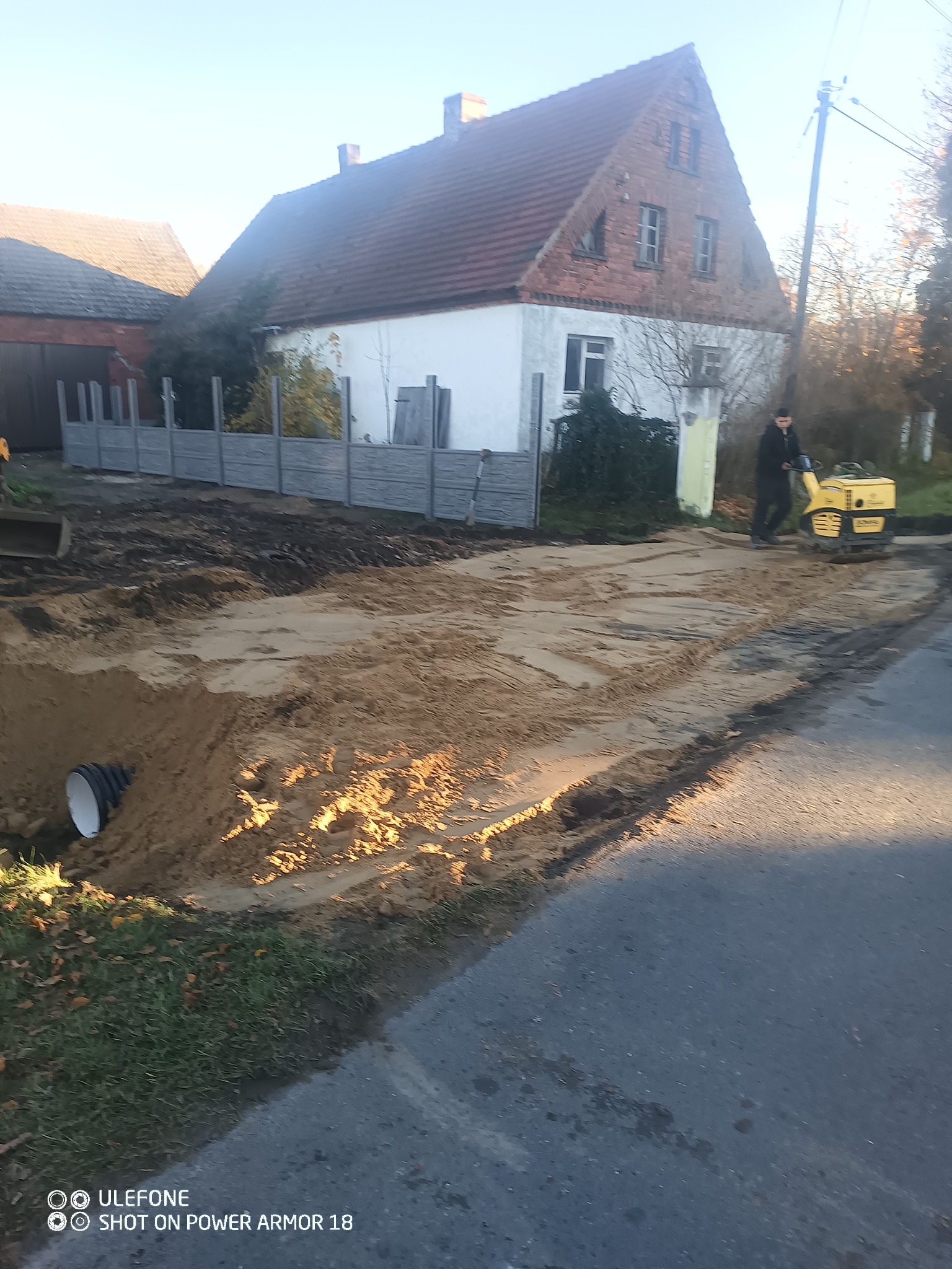 Prace ziemne: ubijanie piasku zagęszczarką w wykopie pod drogę. W tle dom z czerwonej cegły i szary płot betonowy. Widoczna rura drenażowa.