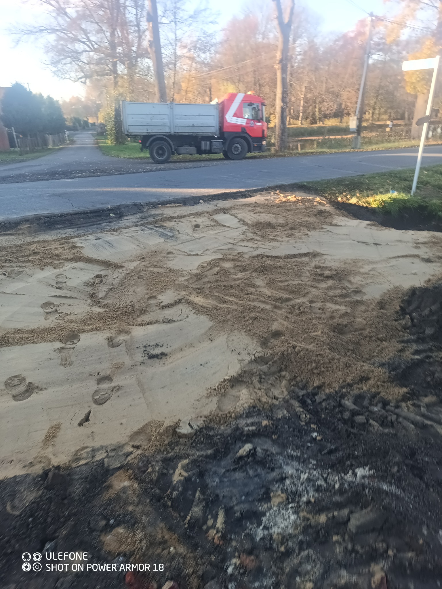 Świeżo nawieziony piasek na placu budowy z widocznymi śladami butów, w tle czerwono-szara wywrotka i drzewa. Utwardzanie terenu pod budowę.