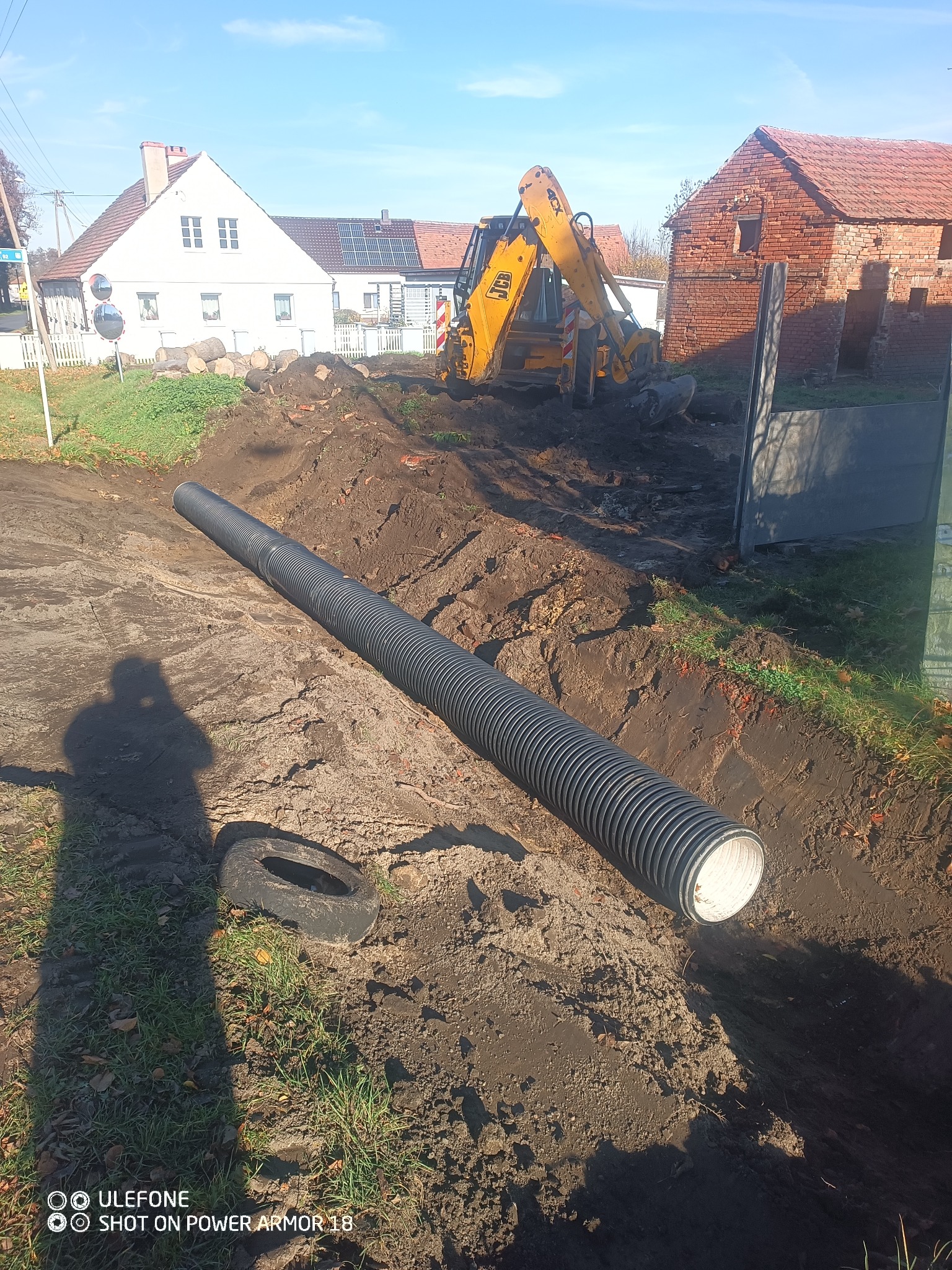 Długa, czarna rura w wykopie ziemnym, prawdopodobnie drenażowa. W tle koparka JCB i zabudowania wiejskie w słoneczny dzień. Cień osoby robiącej zdjęcie.