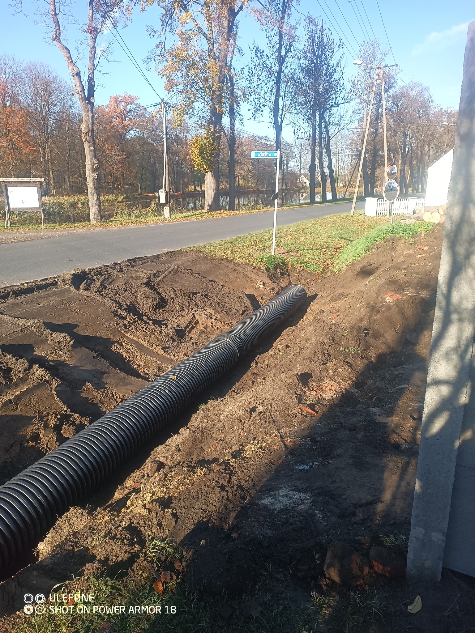 Czarna, karbowana rura ułożona w wykopie ziemnym wzdłuż drogi. W tle drzewa i znak drogowy. Widoczny fragment betonowego elementu. Słoneczny dzień.