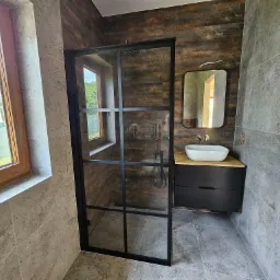 Nowoczesna łazienka z szarymi płytkami imitującymi kamień, kabiną prysznicową typu walk-in z czarnymi profilami, umywalką nablatową na drewnianym blacie i czarną szafką pod umywalką oraz lustrem...