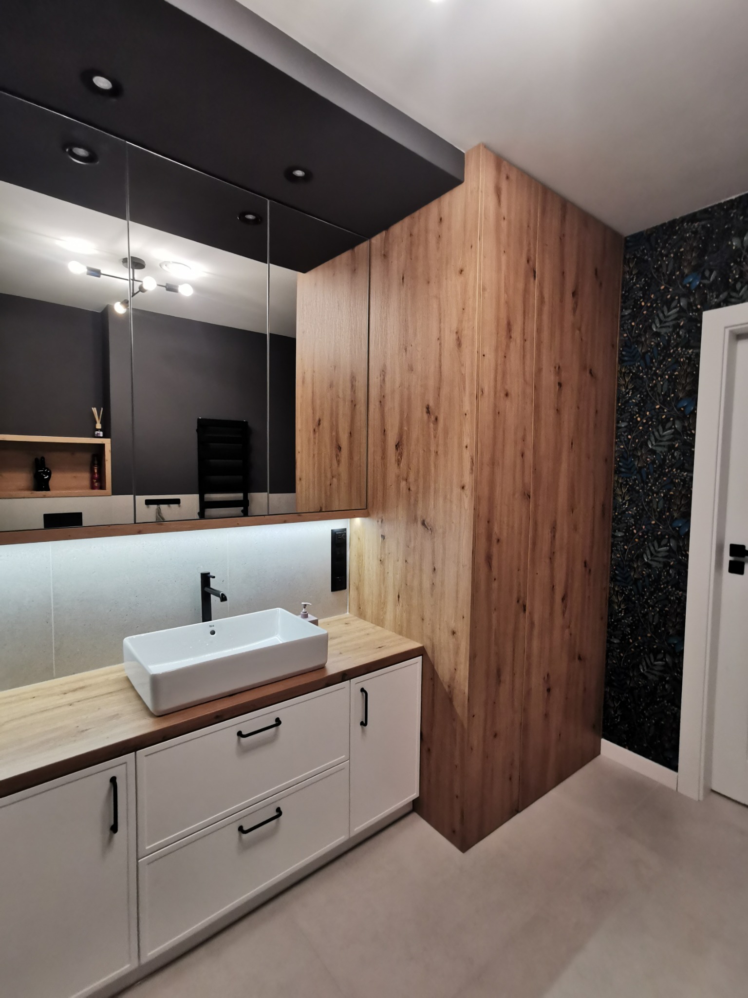 Nowoczesna łazienka z drewnianymi akcentami: szafka pod umywalkę z białymi frontami i czarnymi uchwytami, umywalka nablatowa, lustro z podświetleniem LED, drewniana zabudowa ściany i tapeta...