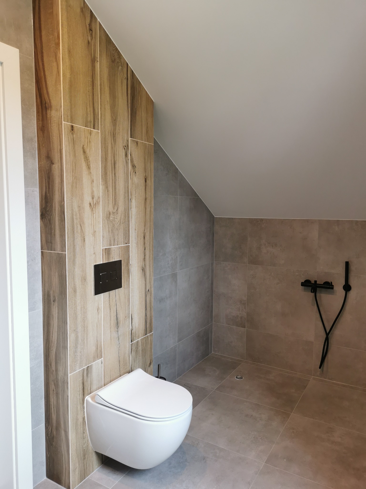 Nowoczesna łazienka na poddaszu z drewnopodobnymi i szarymi płytkami, wisząca toaleta z czarnym przyciskiem, czarna armatura prysznicowa.
