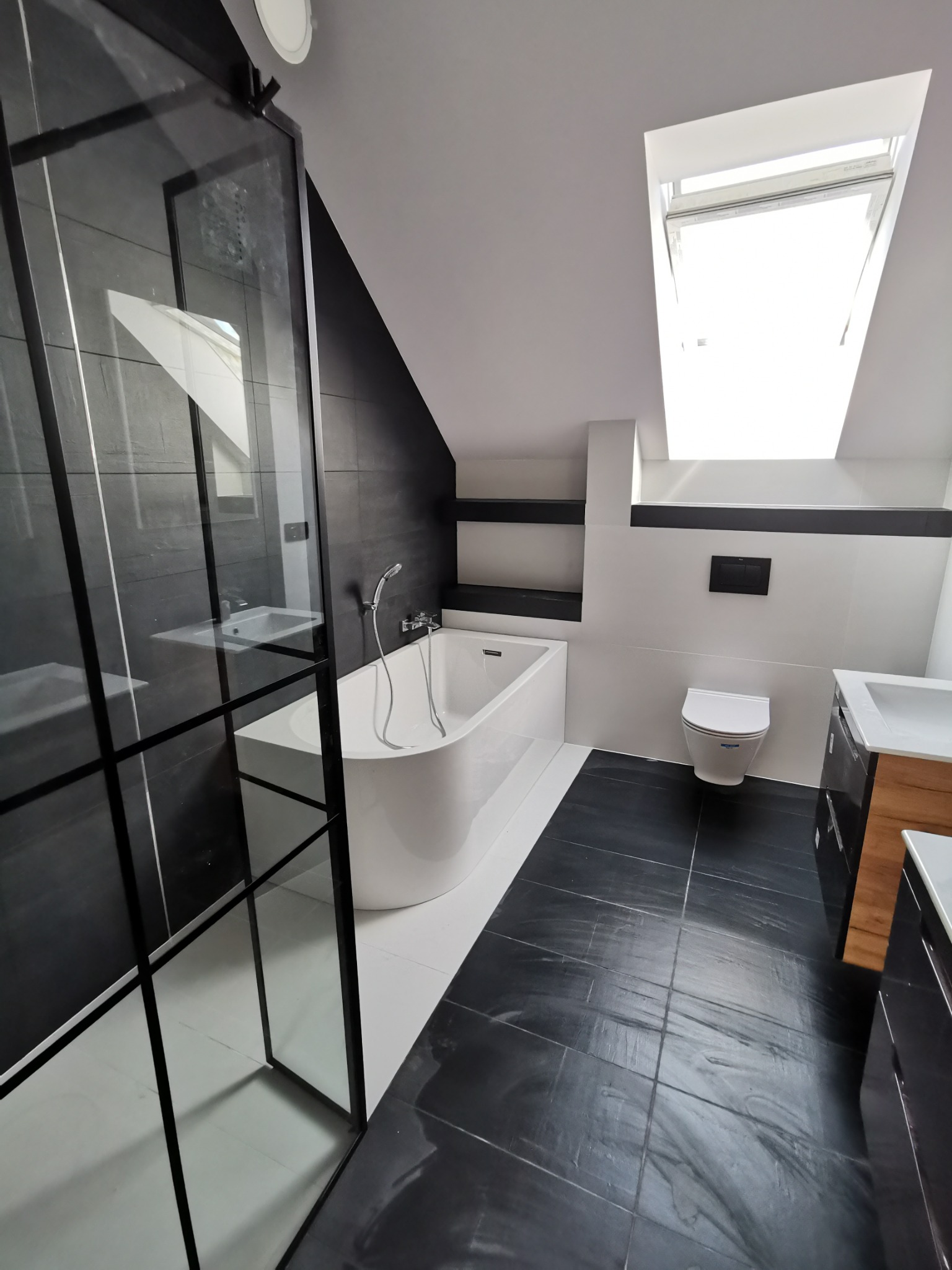 Nowoczesna łazienka na poddaszu z czarno-białymi płytkami, wolnostojącą wanną, kabiną prysznicową typu walk-in z czarnymi profilami, toaletą i szafką z umywalką z drewnianymi elementami.