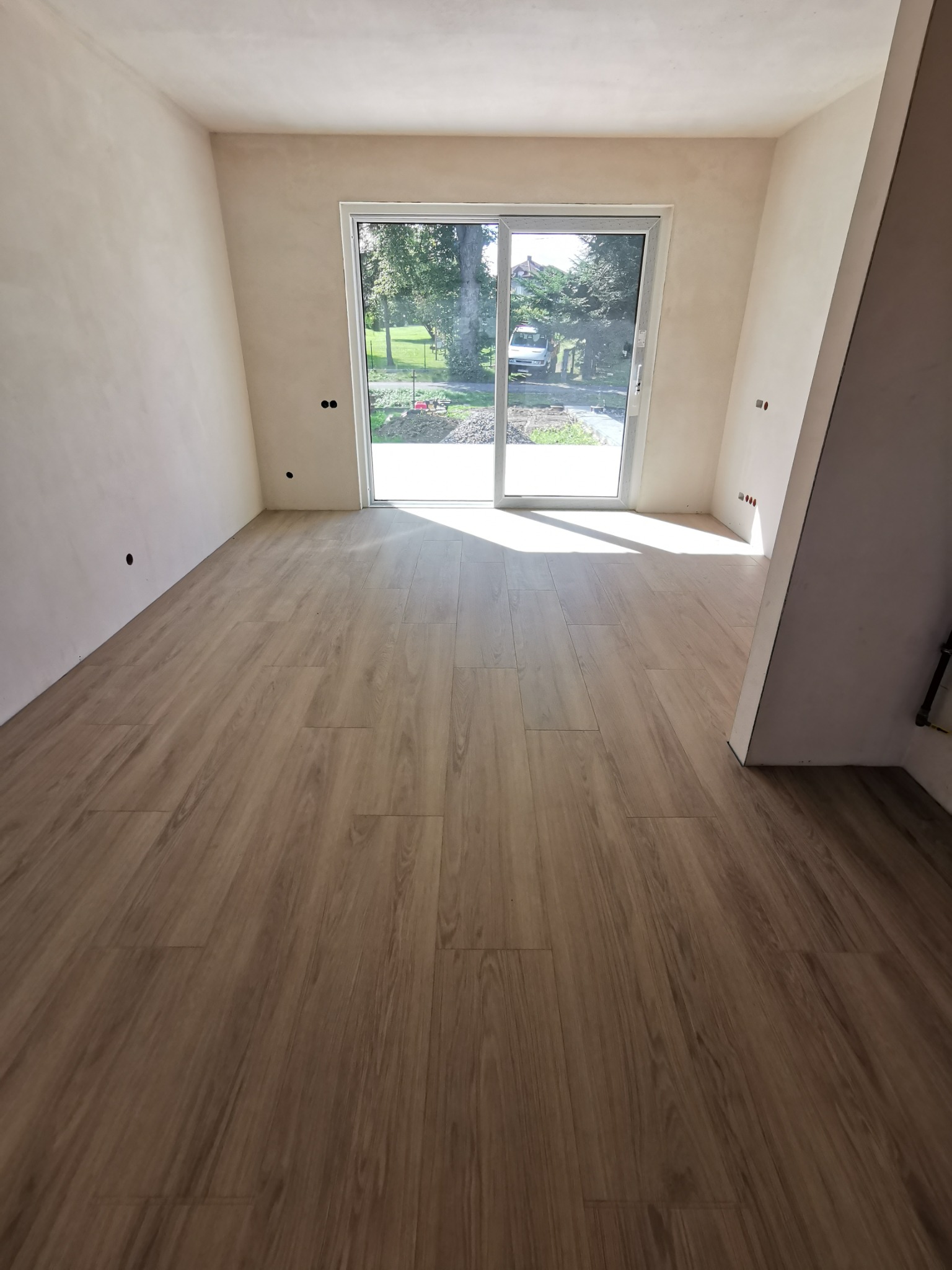 Wykończone wnętrze z drewnianą podłogą, surowe ściany przygotowane do malowania i duże okno balkonowe z widokiem na zieleń.