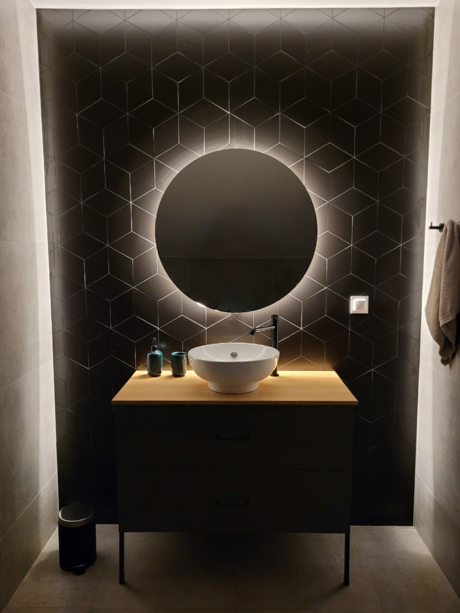 Nowoczesna łazienka z czarnymi heksagonalnymi płytkami, okrągłym lustrem z podświetleniem LED, białą umywalką nablatową i drewnianym blatem szafki, uzupełniona o dyskretny kosz na śmieci...