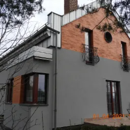 Dwukondygnacyjny dom z szarą elewacją i ceglaną górną częścią, z balkonami i okrągłym oknem na poddaszu, sfotografowany z dołu przez gałęzie drzew.