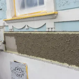 Pracownik w białym kombinezonie i kapeluszu nakłada szarą, strukturalną masę na elewację budynku z niebieskimi elementami dekoracyjnymi, zabezpieczoną taśmą malarską wokół okna.