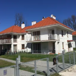 Nowoczesne, białe budynki z czerwonymi dachami krytymi dachówką, ogrodzone metalowym płotem, z numerem domu 109 na elewacji, widoczne w słoneczny dzień.