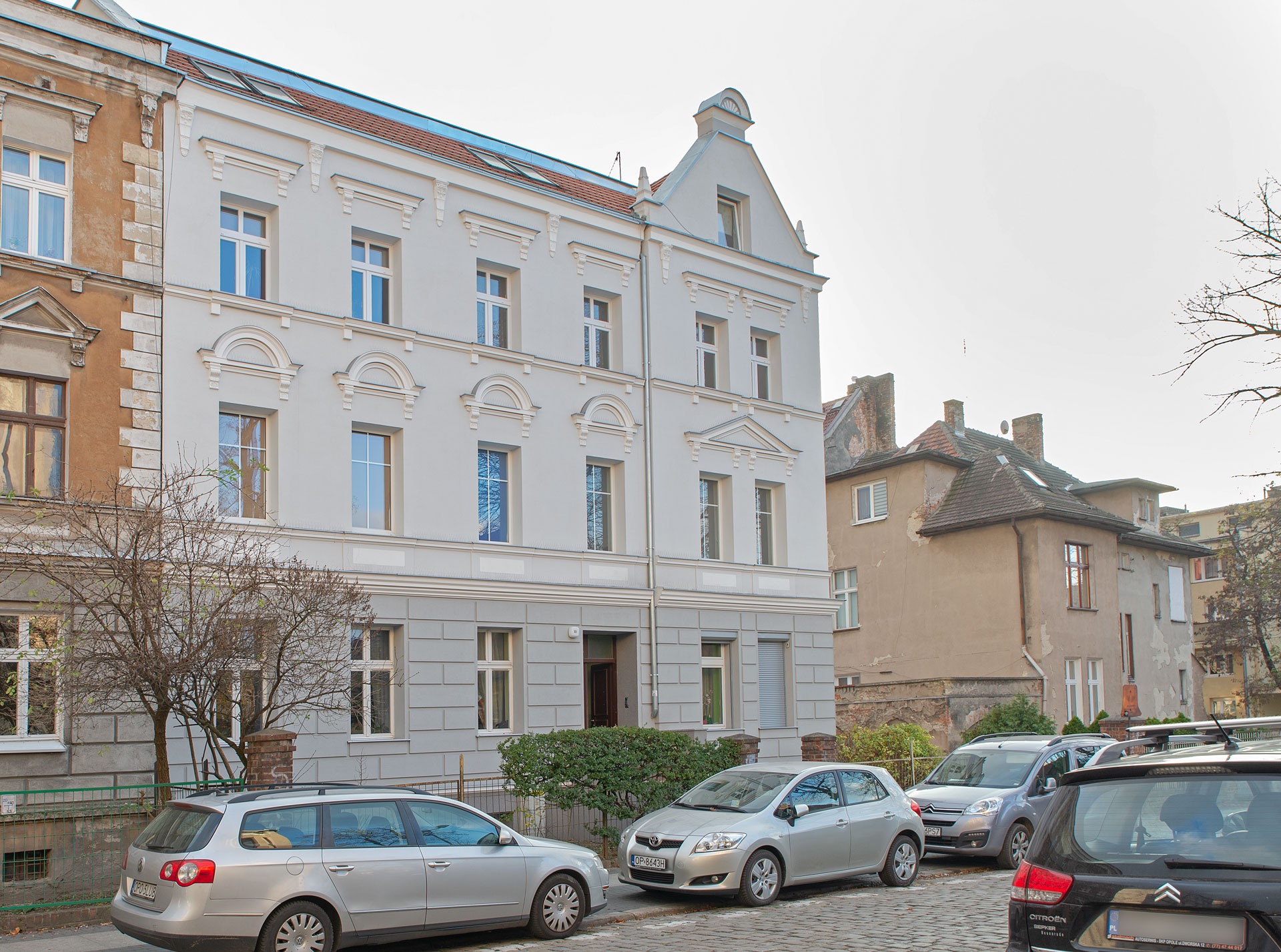 Odnowiona kamienica z szarą elewacją i białymi detalami architektonicznymi, widoczna z perspektywy ulicy z zaparkowanymi samochodami i fragmentem sąsiedniej, starszej kamienicy.