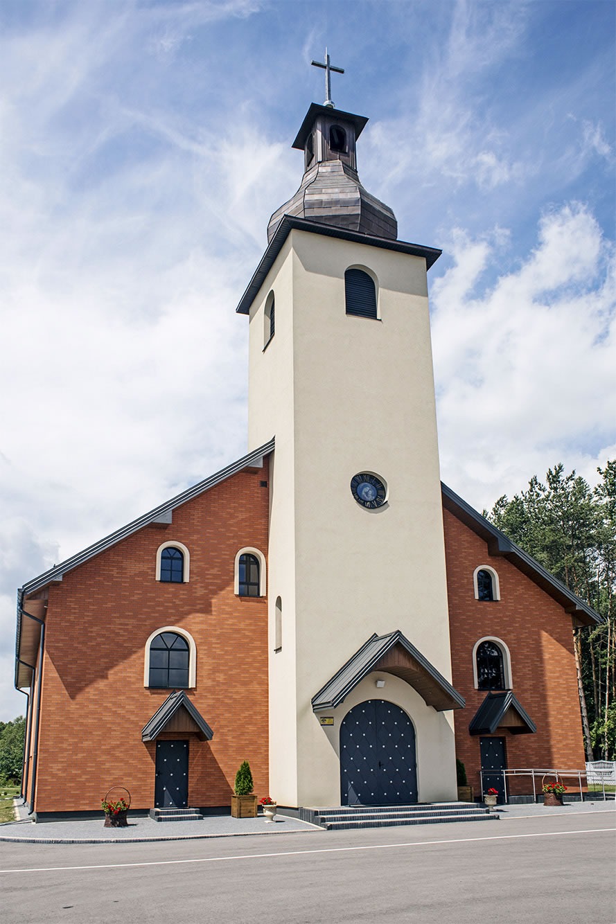 Nowoczesny kościół z ceglaną elewacją i wysoką, kremową wieżą zwieńczoną krzyżem, widoczne detale architektoniczne, takie jak okrągły zegar na wieży i ciemne, łukowe drzwi wejściowe.